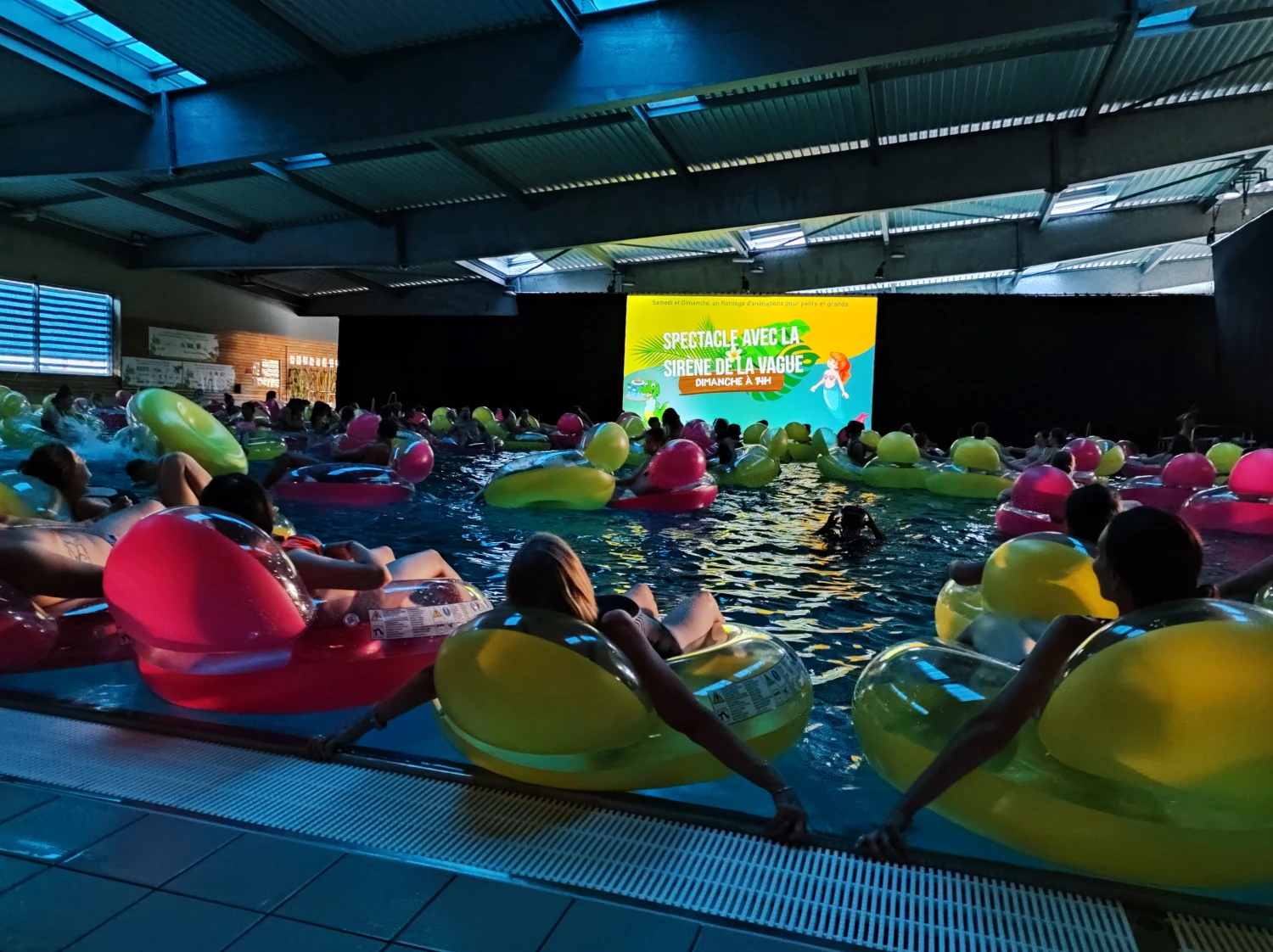 Photo prise dans une piscine plongée dans le noir. Un écran géant est placé au centre de la photo. Le bassin est remplit de personnes sur des boudins flottant en regardant la scéance.